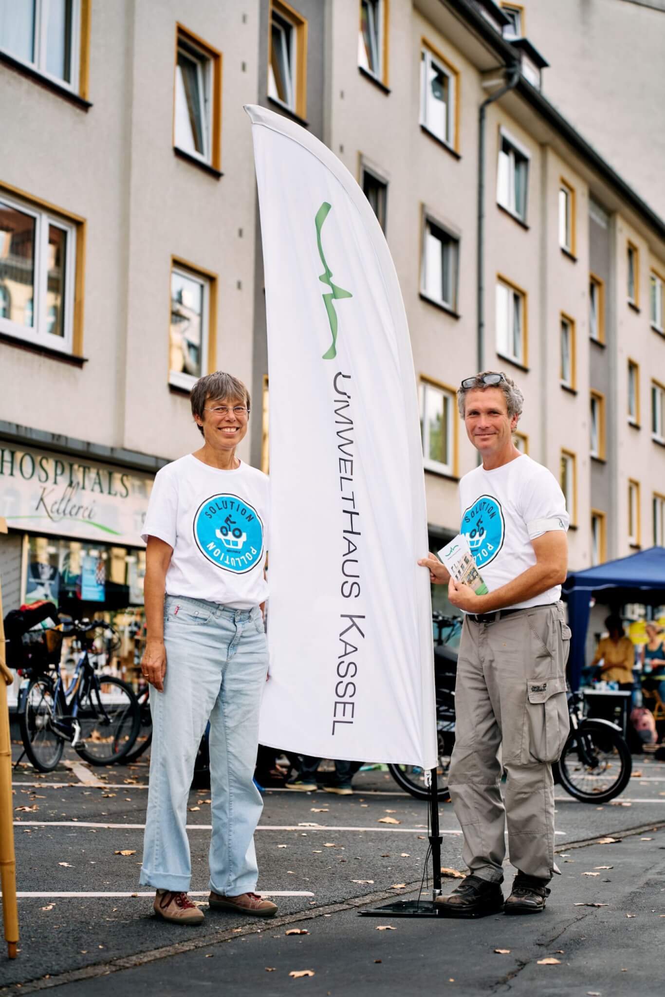Eine Frau und ein Mann in weißen „Solution Pollution“ T-shirts stehen lächelnd neben einer „Umwelthaus Kassel“ Beachflag im Freien und repräsentieren die grüne Zusammenarbeit in Kassel.