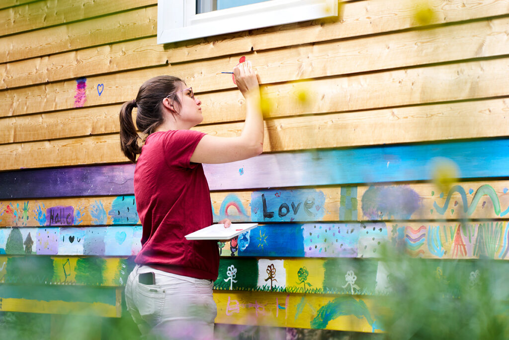 Eine junge Frau bemalt konzentriert die Holzfassade eines Gebäudes mit bunten Farben und Symbolen; das Bild steht für kreatives Engagement und die gemeinschaftliche Gestaltung des urbanen Raums.