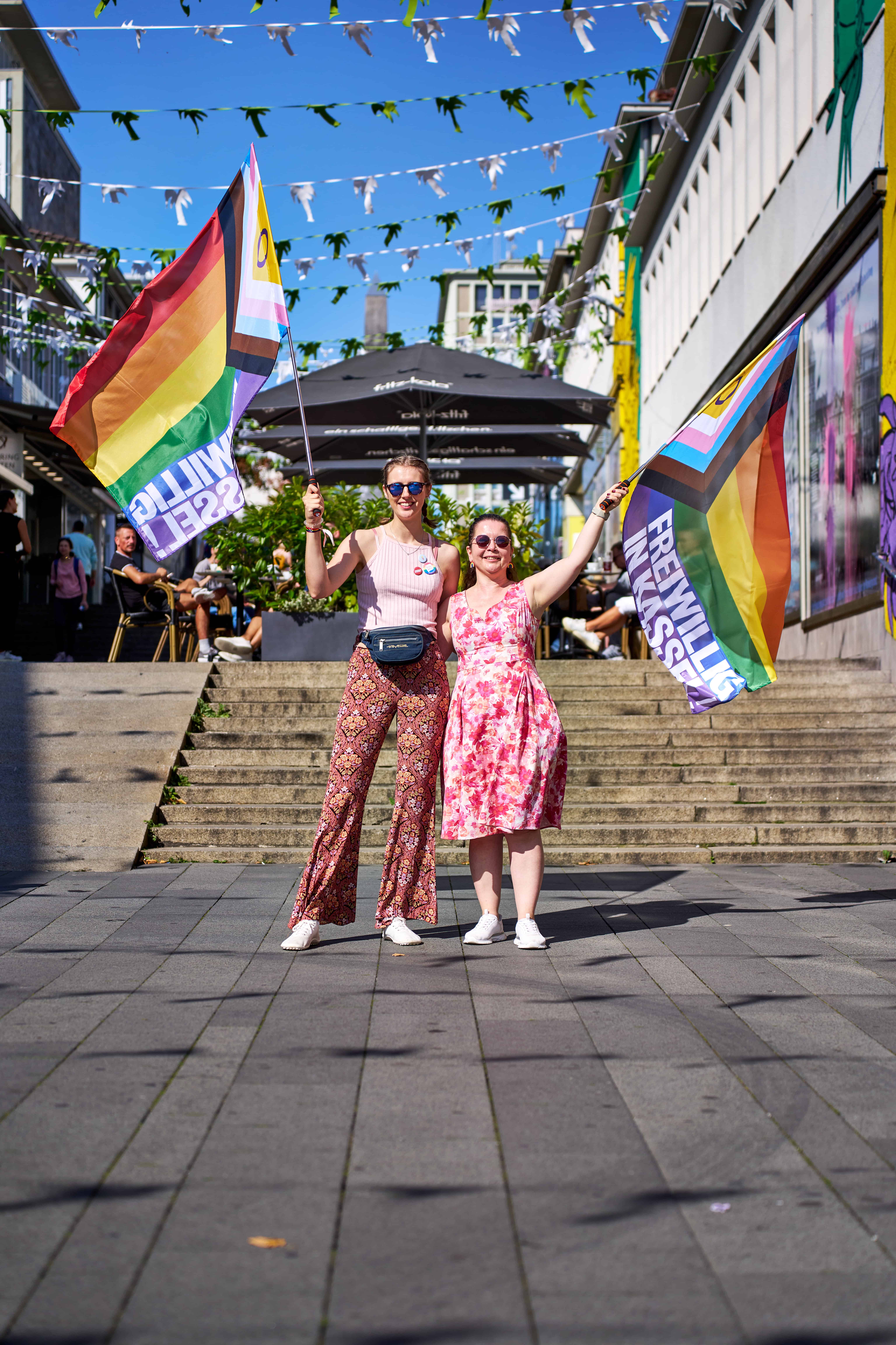 Eine Ganzkörperaufnahme von zwei Frauen im Freien, die auf einer gepflasterten Fußgängerzone vor einer breiten Steintreppe stehen. Beide Frauen lächeln und halten große Progress-Pride-Flaggen hoch, auf denen der Text „FREIWILLIG IN KASSEL!“ zu lesen ist. Die Frau links trägt ein rosa geripptes Tanktop und eine gemusterte Schlaghose, die Frau rechts ein rosa-weiß geblümtes Kleid. Über ihnen hängen dekorative weiß-grüne Wimpelketten quer über die Straße. Im Hintergrund sind Straßencafés unter schwarzen Sonnenschirmen und eine Stadtstraße unter einem strahlend blauen Himmel zu sehen.