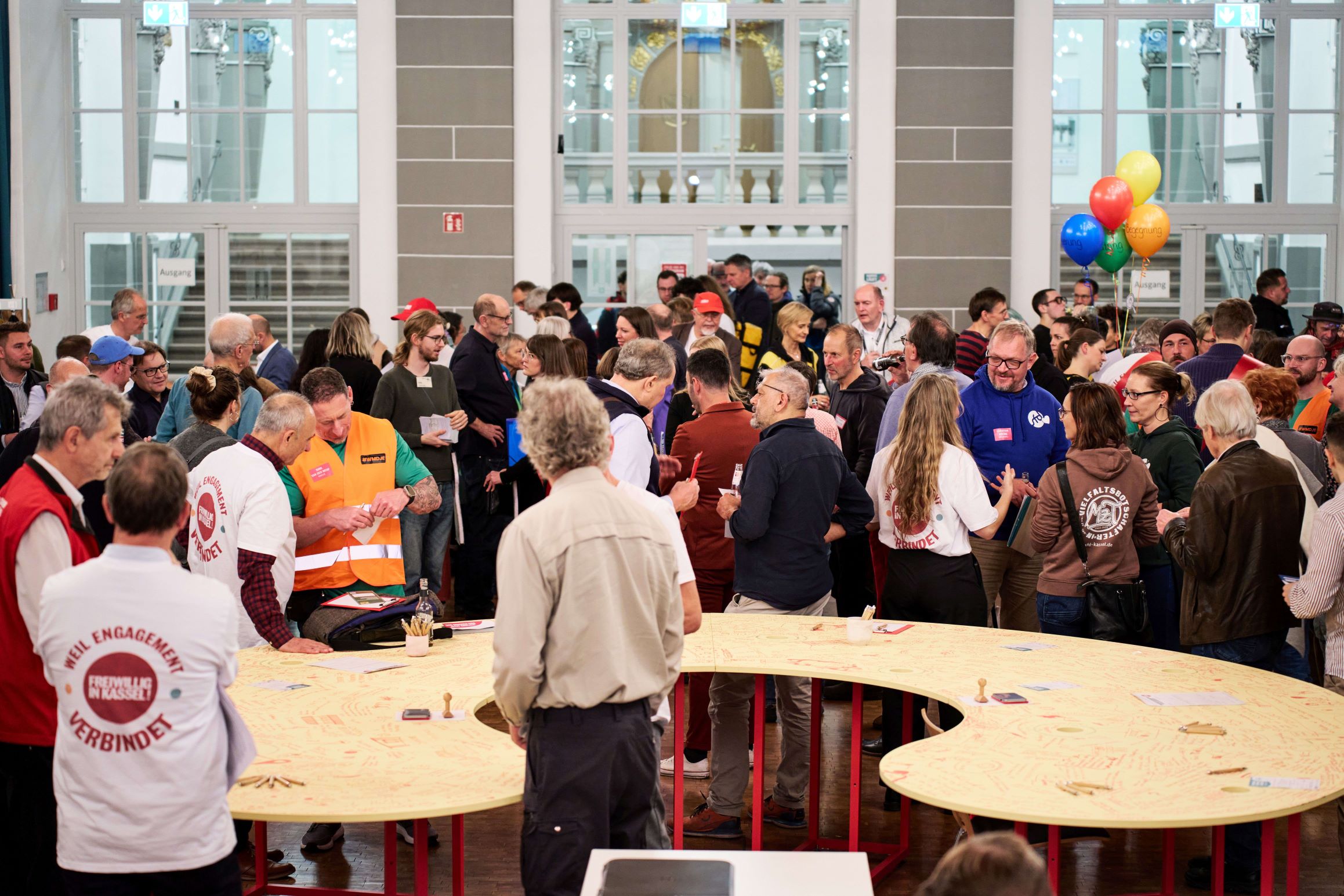 Eine belebte Netzwerkszene im Bürgersaal Kassel; zahlreiche Vertreter aus Wirtschaft und Ehrenamt stehen an geschwungenen gelben Tischen im Dialog, um gemeinsame soziale Projekte und Kooperationen zu vereinbaren.