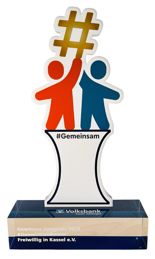 Preis-Skulptur des Jurypreises 2025 „#GemeinsamBesser“. Zwei stilisierte Figuren in Rot und Blau stehen nebeneinander und halten gemeinsam ein goldenes Hashtag-Zeichen hoch. Auf dem Sockel steht „Gewinner Jurypreis 2025 #GemeinsamBesser – Freiwillig in Kassel e. V.“ mit Logo der Volksbank Kassel Göttingen.