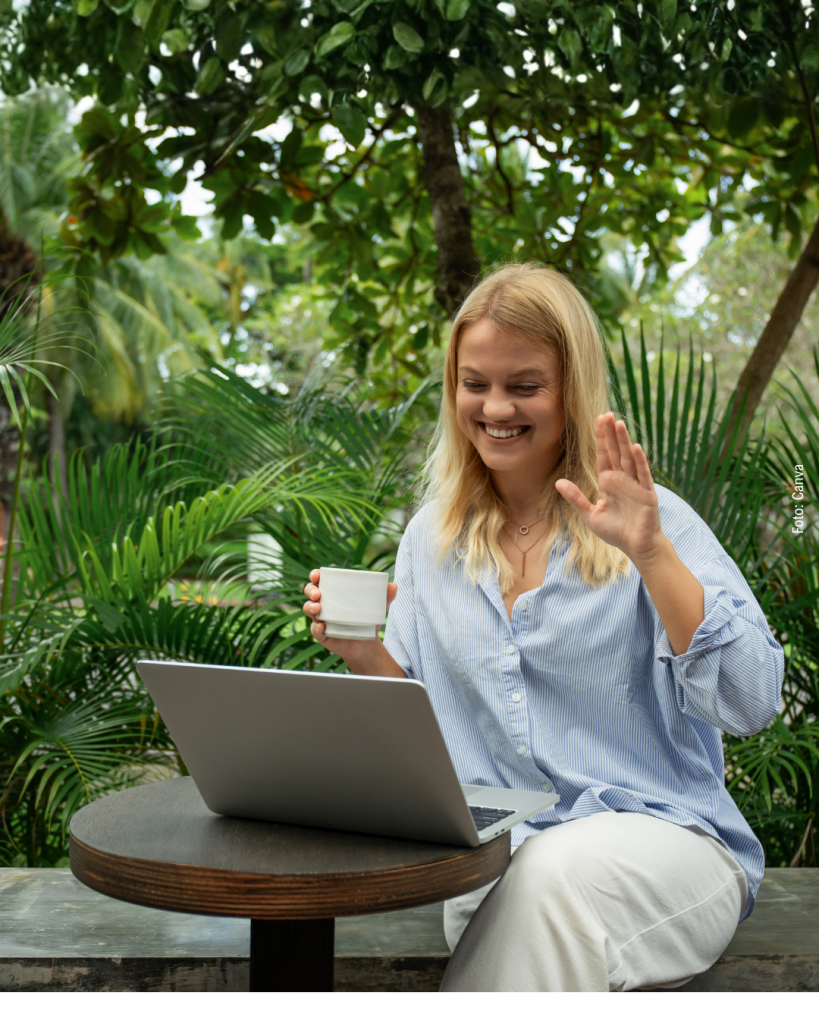 Eine blonde Frau mit Bluse sitzt vor grünen Pflanzen und winkt in den aufgeklappten Laptop der vor ihr steht.